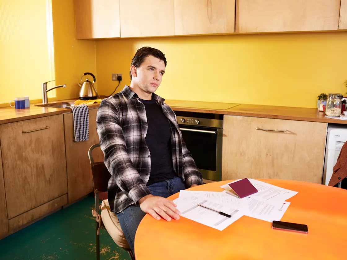 Un hombre sentado a la mesa de su cocina, con documentos y un pasaporte extranjero extendidos delante.